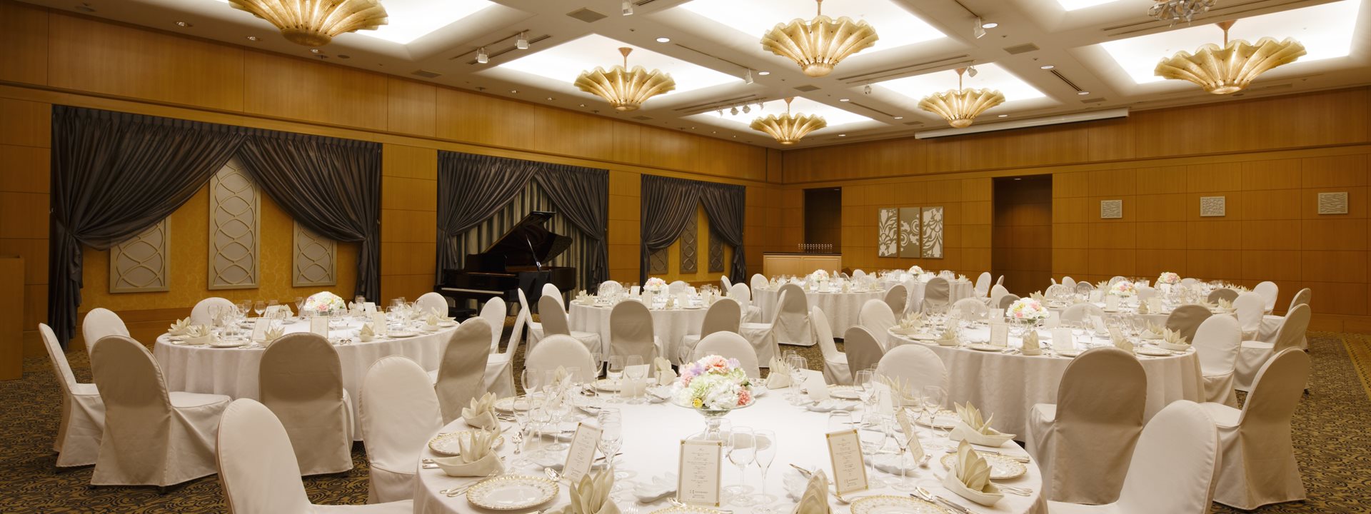 This is the "Ambassadors Ballroom", a medium-sized banquet hall located in the Yokohama Bay Hotel Tokyu. It features round tables for formal dinners, and a grand piano is positioned at the back.